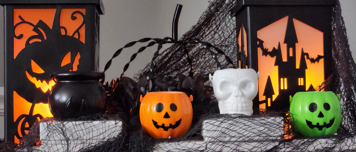 Halloween-themed tabletop decor arrangement featuring two black lanterns with glowing orange jack-o'-lantern and haunted-house cutouts, a black cauldron, a small orange pumpkin, a white skull-shaped container, and a green pumpkin, all draped with black netting.